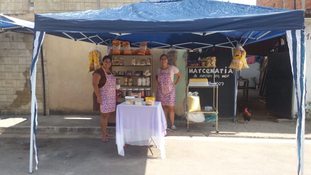 Patrícia e Beatriz, de avental, na barraca de toldo azul com prateleiras de produtos, no dia da inauguração